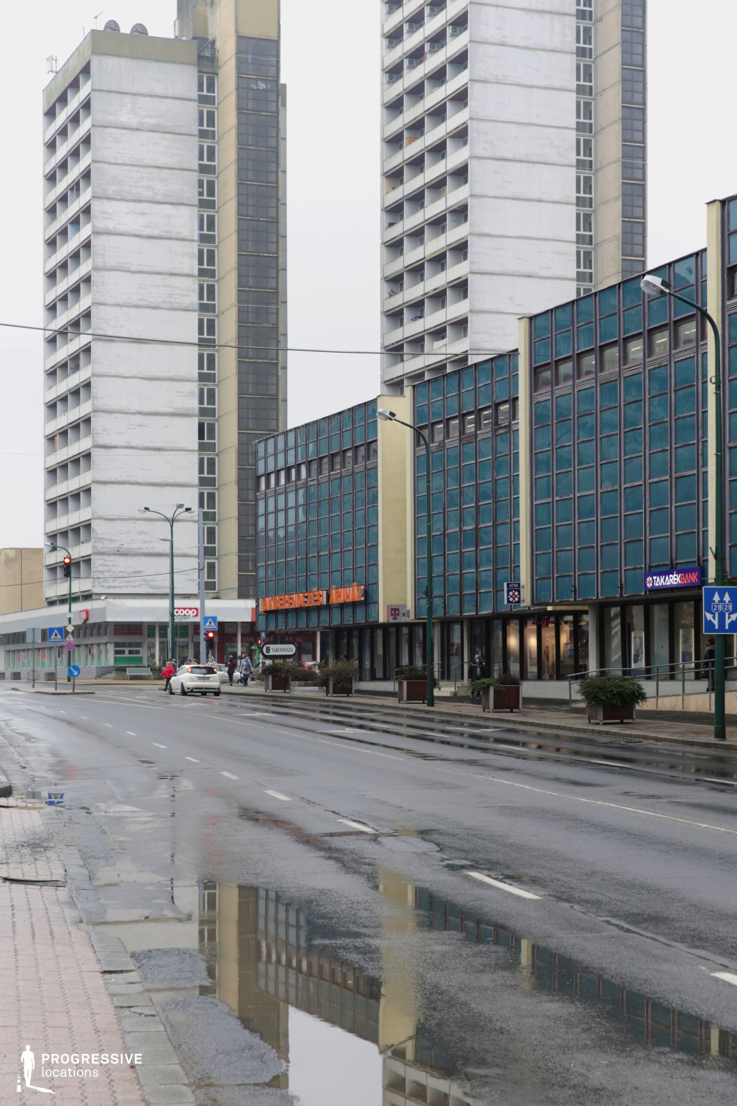 This streetscape showcases a strong juxtaposition between monumental residential high-rises and mid-century office architecture. The tall, concrete towers are stark and symmetrical, while the adjacent commercial building features a striking grid of blue glass panels framed in metal, offering a rhythmic, repetitive aesthetic. The road flanked by wide pavements and evenly spaced lamp posts creates a clean visual corridor with long sightlines, suitable for dynamic tracking shots. The verticality of the high-rises contrasts effectively with the horizontal expanse of the glass-fronted block, enhancing the depth and urban scale of the composition. This location suits film genres such as political drama, cold war-era period films, or minimalist dystopian narratives, and is well-suited for establishing shots, street-level sequences, or introspective urban moments.