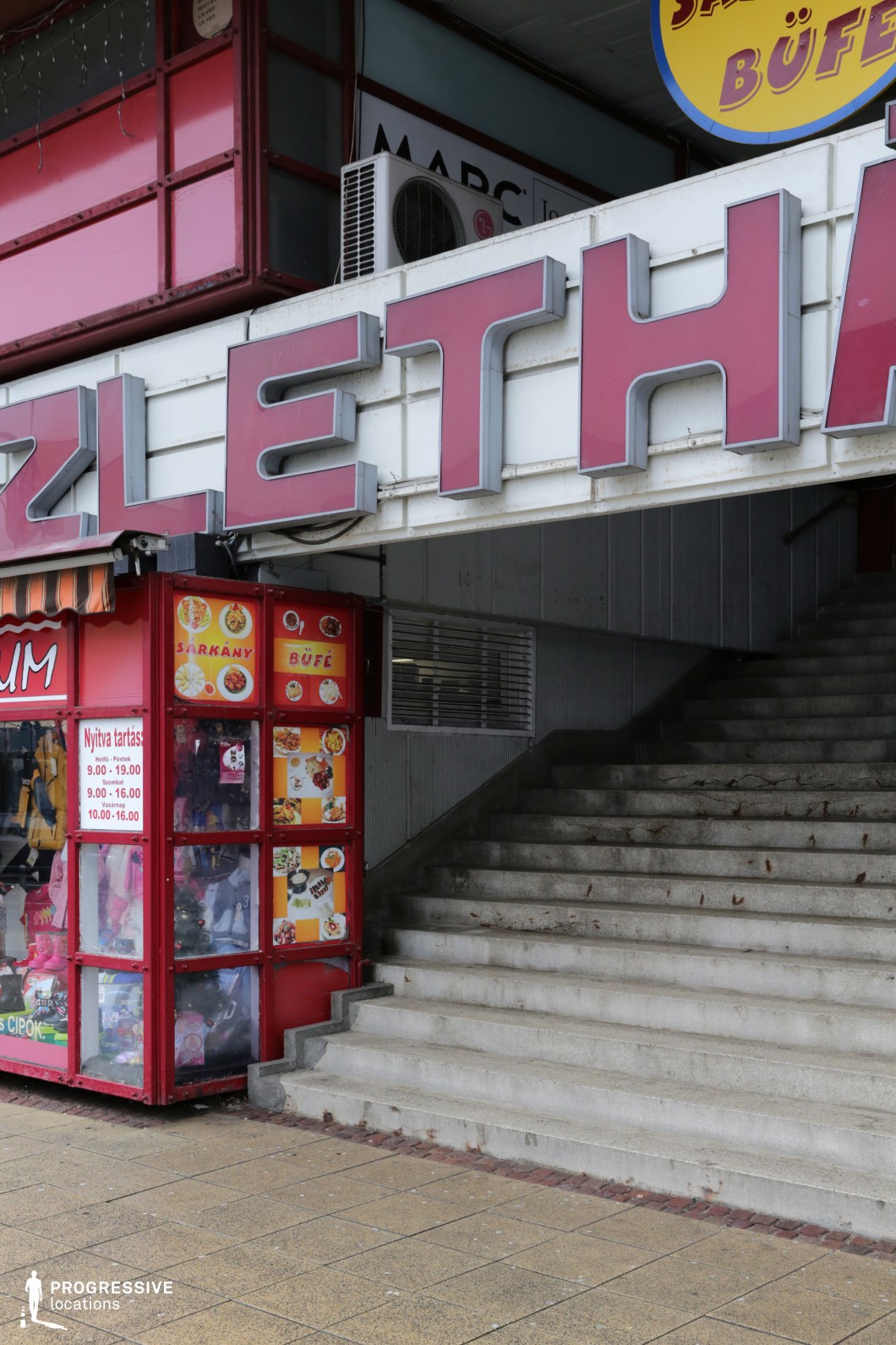 This urban location features a wide concrete staircase leading into a covered walkway beneath a building facade characterized by large, bold signage with a distinctly retro aesthetic. The dominant colors are muted grays and whites, punctuated by vivid red accents on the storefront frames and illuminated lettering, evoking a socialist-era commercial design. The materials include exposed concrete, metal paneling, and transparent plastic windows, contributing to a utilitarian and slightly worn look. The low-angle perspective highlights the geometric rhythm of the steps and signage, creating a strong compositional focus. Ideal for productions seeking vintage Eastern European cityscapes, this location supports filming in genres like social realism, drama, or retro-themed narratives, particularly for street-level scenes, commercial areas, or transitional urban spaces.