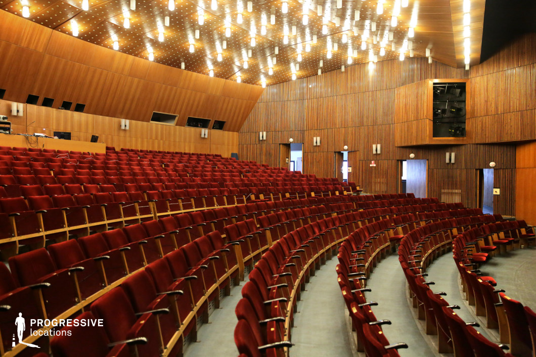 This large auditorium features a sweeping, curved seating arrangement composed of plush red seats with wooden armrests, creating a warm and unified aesthetic. The walls and ceiling are fully clad in vertical and angled wood panels, contributing to excellent acoustics and a cohesive visual identity. A dense grid of suspended cylindrical light fixtures provides even illumination while enhancing the architectural rhythm of the space. The structure incorporates built-in technical booths, recessed wall lighting, and minimal decorative elements, emphasizing functionality and modernist simplicity. With its spacious layout, unobstructed sightlines, and cinematic symmetry, this location is ideal for filming scenes set in lecture halls, performance venues, or institutional settings, particularly for dramas, political thrillers, or historical narratives.