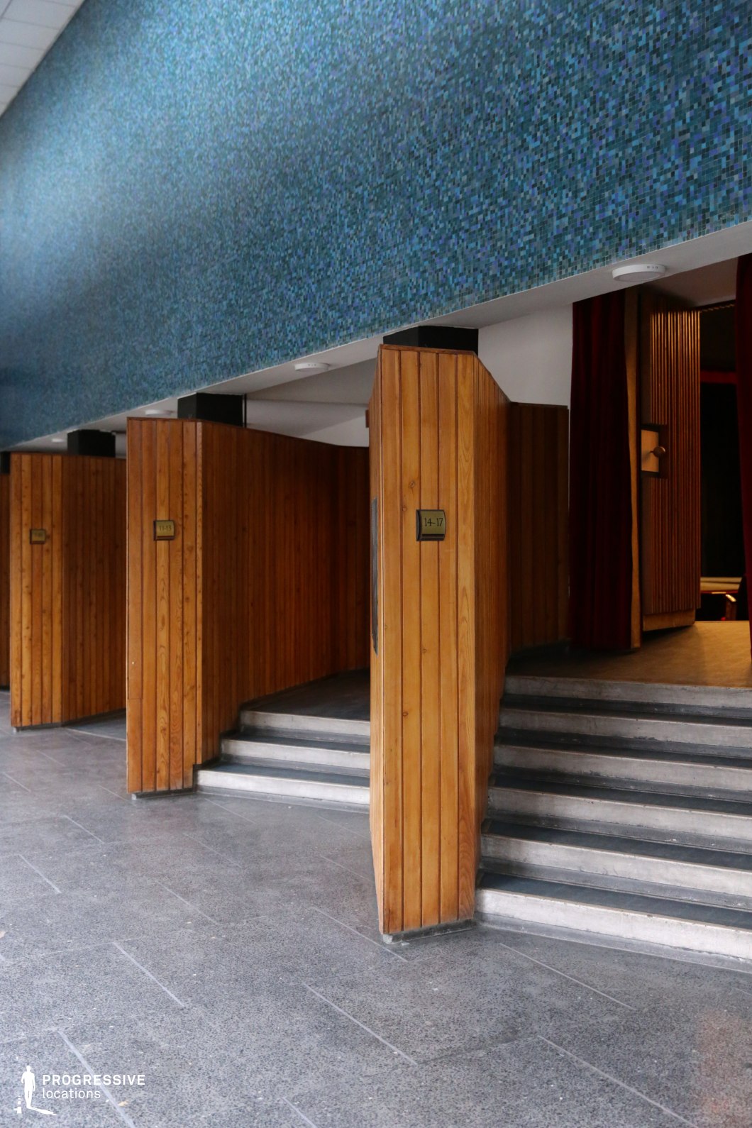 This location showcases a distinctive mid-century modern interior defined by vertical wooden paneling and a bold blue mosaic tile wall above. The entrance pods are separated by angular wooden structures leading to a staircase, each labeled with signage indicating seating sections, suggesting the space is part of a theater or auditorium. The flooring consists of dark grey polished stone, adding a durable, institutional feel, while the warm-toned wood contrasts with the cool blue hues above, creating a balanced yet visually engaging palette. The geometric forms of the stairs, walls, and overhead elements lend the location a structured, cinematic quality. This setting is particularly suitable for filming retro-inspired productions, institutional environments, or transitional interior scenes in drama, period pieces, or mystery genres due to its strong visual identity and architectural character.