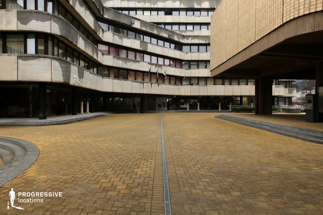 This urban courtyard is framed by a Brutalist-style office complex with a distinctive curved concrete facade, featuring repetitive window patterns and weathered exterior surfaces. The yellow-toned brick paving, intersected by a central drainage channel, contrasts with the raw concrete structures. An overhanging section with textured tile cladding adds depth and an imposing presence to the scene. The harsh geometry and institutional aesthetic of the space make it an ideal setting for dystopian, political thriller, or neo-noir films, evoking themes of bureaucracy, surveillance, or corporate power struggles. Its stark atmosphere lends itself to tense, introspective, or dramatic storytelling.