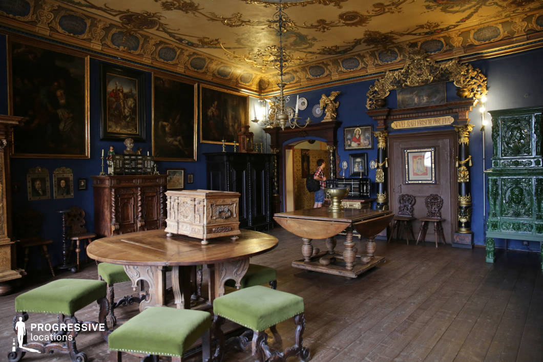 This richly decorated interior showcases an opulent historical room with deep blue walls adorned by large, gold-framed oil paintings and baroque architectural ornamentation. The elaborately carved wooden furniture, including circular and rectangular tables with green velvet stools, adds a Renaissance-era aesthetic. The ornate ceiling features gold-painted motifs and reliefs, contributing to the room's regal character. Textural contrast is provided by the dark wood floor and a striking, glossy green ceramic tile stove. The spatial proportions are moderately generous, with enough depth for dynamic camera movement, while the symmetrical arrangement of wall elements enhances compositional balance. This location is ideal for filming period dramas, historical reconstructions, or artistic documentaries, particularly those requiring a noble or museum-like interior with strong visual detail and old-world atmosphere.