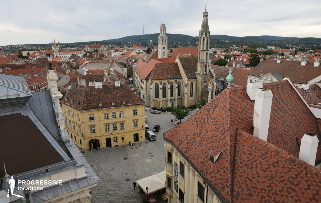 This elevated view captures a densely built, historic European town characterized by a cohesive array of red-tiled rooftops, cobblestone streets, and prominent ecclesiastical architecture. The focal point is a central square bordered by baroque and gothic-revival buildings, including a church with a soaring, ornate tower and lancet windows. The architecture combines soft yellows, earth tones, and stone textures, contributing to a harmonious and authentic old-town aesthetic. The layered rooflines, winding alleys, and varied architectural styles provide rich visual depth and historical continuity. This location is highly suitable for period dramas, historical reconstructions, or travel-themed productions requiring panoramic establishing shots of a preserved heritage setting.