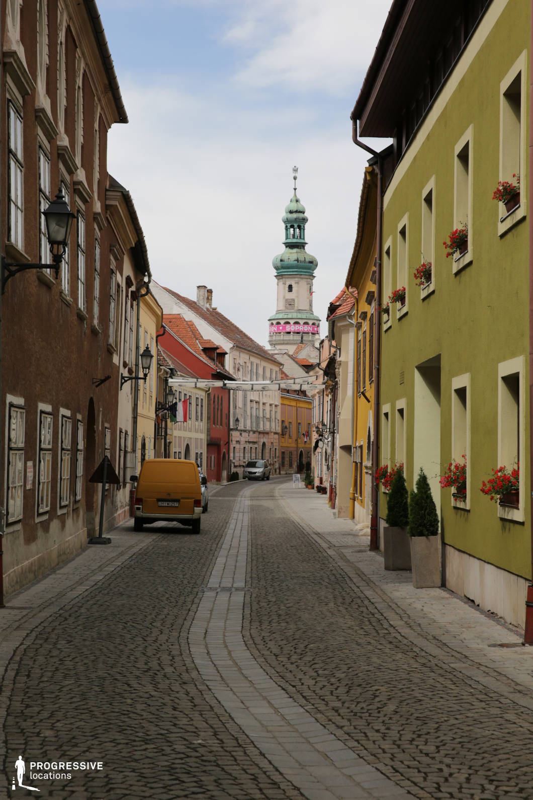 This narrow, curving cobblestone street is framed by a mix of well-preserved, colorful historic buildings with varying facades in earthy browns, muted yellows, and vibrant greens, creating a harmonious yet dynamic visual rhythm. The textures of stucco, stone, and painted plaster highlight the old-world character, complemented by traditional window shutters and wrought-iron lamp posts. A dominant architectural focal point is the ornate Baroque-style tower rising in the distance, its green patinated dome and tiered spire drawing the eye upward and anchoring the composition. The street's scale is intimate, with two- to three-story buildings tightly enclosing the space, enhancing a sense of enclosure and depth. The visual continuity of the curved paving stones, symmetrical window arrangements, and the central perspective make this location ideal for period films, historical dramas, or romantic city scenes requiring an authentic European ambiance with strong architectural character.