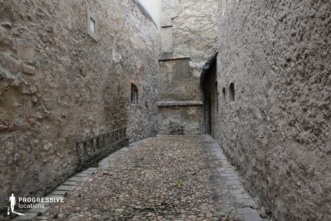 This image showcases a narrow, enclosed alleyway bordered by rough, aged stone walls that evoke a distinctly historical and timeworn atmosphere. The cobblestone pavement is uneven and varied in tone, contributing to the textured, authentic feel of the space. The walls exhibit a range of surface treatments, from exposed bricks to plastered and weathered masonry, all in muted earth tones that enhance the sense of age and seclusion. Small architectural details, such as barred windows and arched recesses, provide subtle depth and character. The confined scale and verticality of the space, combined with the linear perspective captured from a central eye-level viewpoint, reinforce the sense of enclosure. This location is ideal for historical dramas, thrillers, or period films, offering a visually rich backdrop for intense, secretive, or introspective scenes.