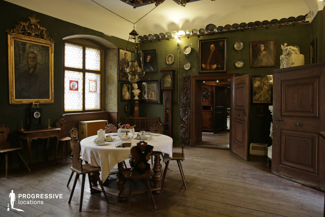 This interior presents a richly detailed historical dining room with dark green walls, wooden flooring, and antique furnishings. A round wooden table with turned legs occupies the center, surrounded by carved wooden chairs, and covered with a white tablecloth and classic tableware. The walls are densely decorated with oil portraits in ornate frames, pewter plates, and intricate wall carvings, creating a gallery-like effect. Decorative elements such as a vintage grandfather clock, a brass hanging lamp, and an arched window with stained glass inserts further enhance the period ambiance. The architectural detailing includes solid wooden doors with paneling and a heavily carved doorframe, adding a sense of craftsmanship and historic authenticity. This visually layered, atmospheric space is ideal for period dramas, historical documentaries, or costume films, particularly scenes requiring a formal, old-world European interior.