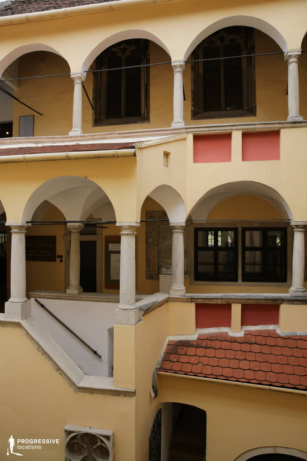 This location features a two-story inner courtyard defined by symmetrical arcades supported by robust stone columns with simple capitals, characteristic of Renaissance architecture. The building's façade is painted in soft ochre tones, accented with muted terracotta panels and warm red roof tiles, establishing a balanced, historical ambiance. The arched windows on the upper level are framed in dark stone, contributing to the vertical rhythm of the composition and reinforcing its classical aesthetic. The enclosed space is vertically layered with staircases, vaulted walkways, and recessed entry points, offering varied depth and strong architectural geometry. This space is ideal for period pieces, historical dramas, or artistic productions requiring a well-preserved, European-style courtyard, with excellent framing options for dialogue-heavy scenes, architectural cutaways, or stylized movement sequences.