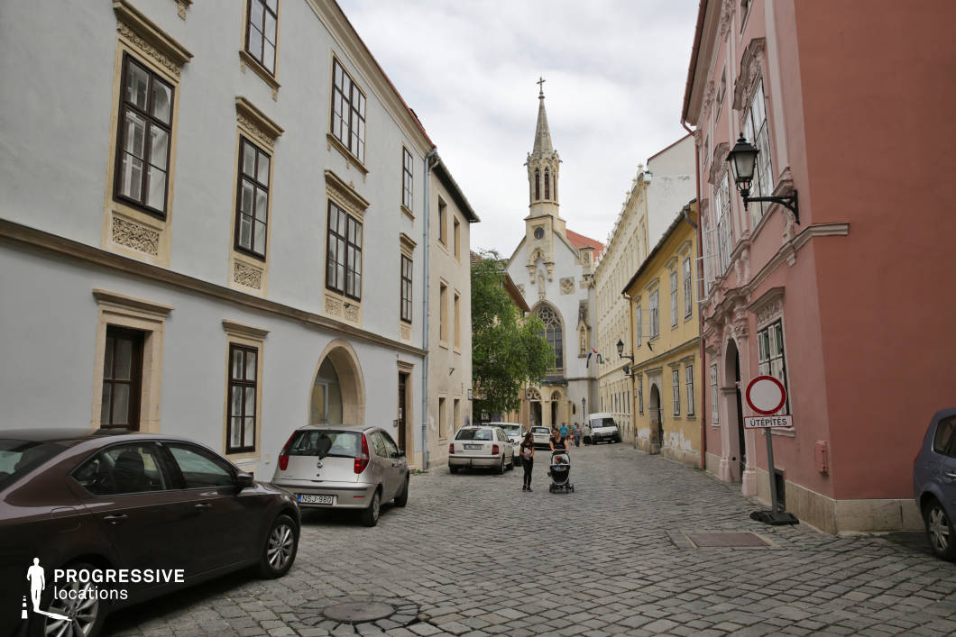 This narrow, cobblestone-paved street is flanked by well-preserved historic buildings featuring classic European architectural styles, including ornate plasterwork, arched doorways, and symmetrically arranged sash windows. The color palette includes muted pastels such as white, beige, pale yellow, and rose pink, contributing to a warm and refined atmosphere. A Gothic Revival church with an elaborately detailed steeple forms the focal point at the end of the street, drawing the eye with its pointed arches and sculpted façade. The street is visually enclosed by the buildings on both sides, creating an intimate, tunnel-like urban environment. The location is ideal for filming scenes that require a period European backdrop, evoking old-town charm and architectural richness, making it suitable for historical dramas, romance films, or art-house projects requiring a quiet, atmospheric city setting.