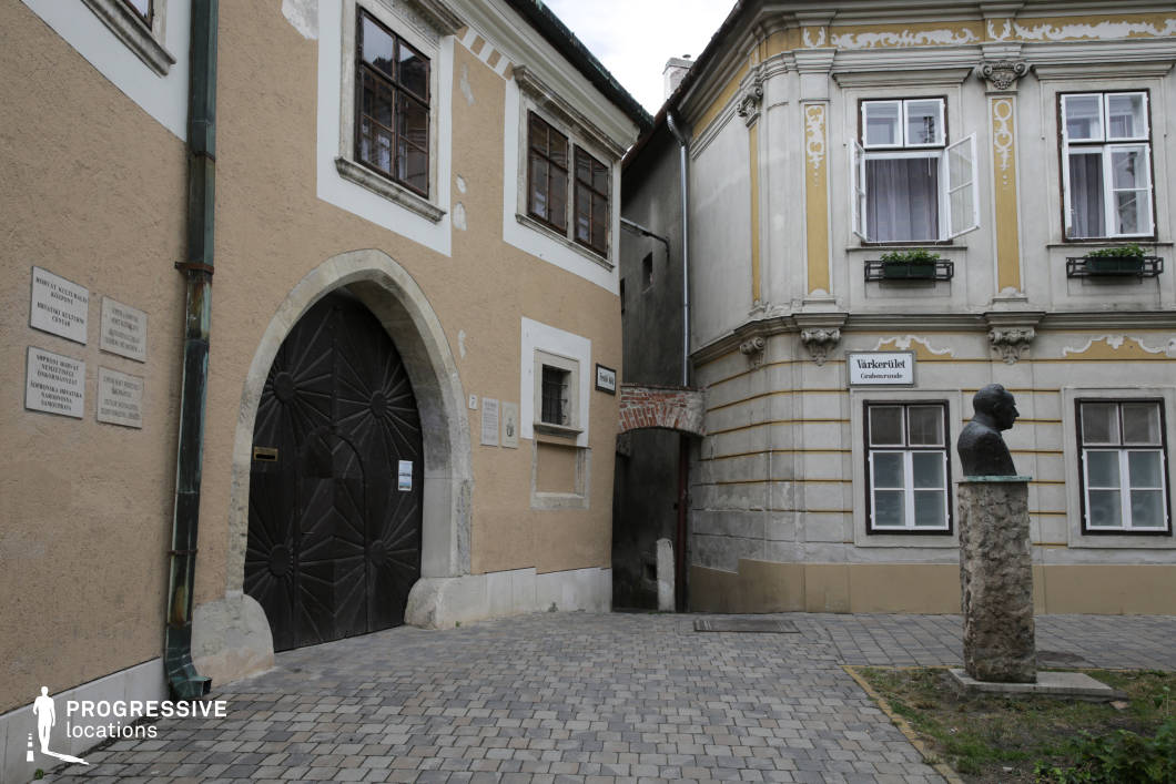 This urban corner features a narrow cobblestone alley framed by two adjoining historic buildings, forming an intimate passage between aged stone facades. On the left, a rustic plastered wall in beige tones houses a large, dark wooden arched door set within a stone frame, adding a medieval touch. The opposing structure showcases Baroque architectural elements, including stucco ornamentation, fluted pilasters, and detailed cornices in muted yellow and grey hues. Windows are symmetrically aligned, some adorned with wrought-iron flower boxes, enhancing the old-world charm. A small bust sculpture on a textured stone pedestal introduces a formal element to the modest plaza. The space is tightly enclosed, with vertical architectural details creating a visually rich, layered backdrop ideal for controlled framing. This location is suitable for filming historical dramas, period pieces, or atmospheric urban scenes that require authentic architectural depth and a confined, characterful setting.