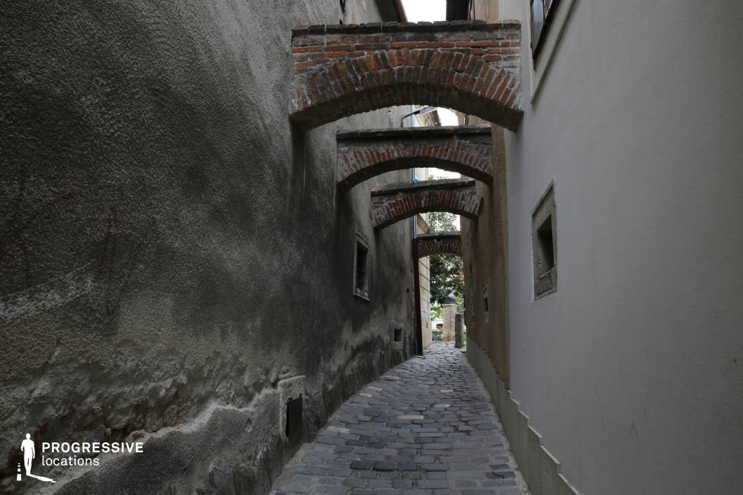 This narrow, cobblestone alleyway is flanked by rough-textured plaster and stucco walls, creating a confined, enclosed spatial experience. A defining architectural feature is the series of evenly spaced, low-slung red brick arches connecting the two buildings overhead, lending a historical and fortified atmosphere to the passage. The muted grey and brown color palette contributes to a worn, timeworn aesthetic, while the curved path subtly directs the eye toward the brighter opening in the distance. The tight proportions and repetitive geometric elements create strong visual rhythm and depth. This location is ideal for filming suspenseful, historical, or dramatic scenes, particularly in genres such as thriller, period drama, or mystery, where atmospheric tension and architectural texture are key visual elements.