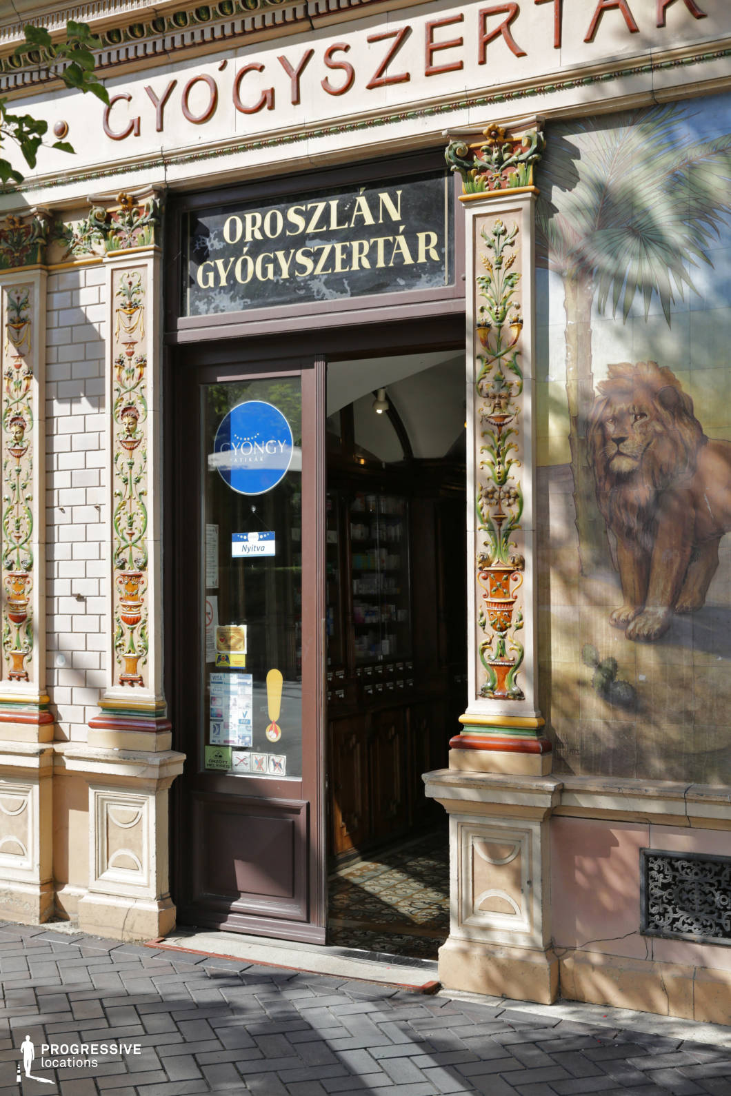 This storefront features a richly decorated historic pharmacy entrance with an elaborate ceramic façade. The design incorporates vertical floral reliefs in vivid greens, yellows, and reds set within cream-toned architectural framing. A large mosaic-style lion mural adds a distinctive and memorable focal point, enhancing the building's unique character. The door is framed by dark wood and glass, offering a glimpse into a traditional interior with wooden cabinetry and patterned tile flooring. The white glazed brickwork and classical cornices further emphasize the late 19th to early 20th century style. The narrow sidewalk in front is paved with dark gray bricks, forming a clean visual base for the colorful elevation. This location is ideal for period productions, commercials, or artistic urban scenes, especially those needing detailed, decorative exteriors with a unique identity and historical ambiance.