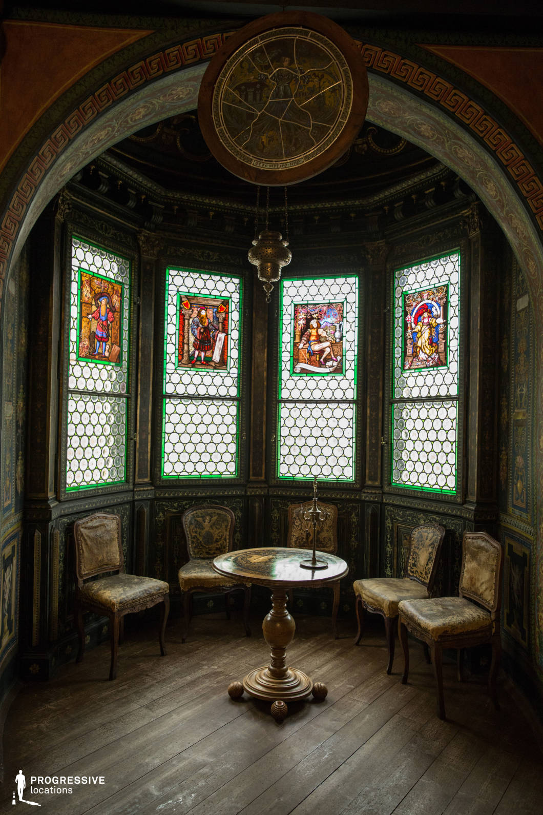 This richly detailed interior features a semi-circular alcove framed by five stained glass windows, each adorned with vivid, classical-style illustrations bordered by green geometric patterns. The dark wood paneling, finely decorated with gold and green ornamental motifs, enhances the historic atmosphere. A circular wooden table with a sculpted pedestal base stands at the center, surrounded by four aged, upholstered chairs in varying conditions, all set atop a wide-planked wooden floor. Above, a hanging pendant lamp and a large circular wall element add vertical dimension and depth. The enclosed, intimate layout and the dominant palette of deep greens, browns, and golds evoke a solemn, timeless elegance. This location is ideal for filming scenes in historical dramas, period pieces, or fantasy genres, particularly those requiring a lavish, introspective, or secretive setting.