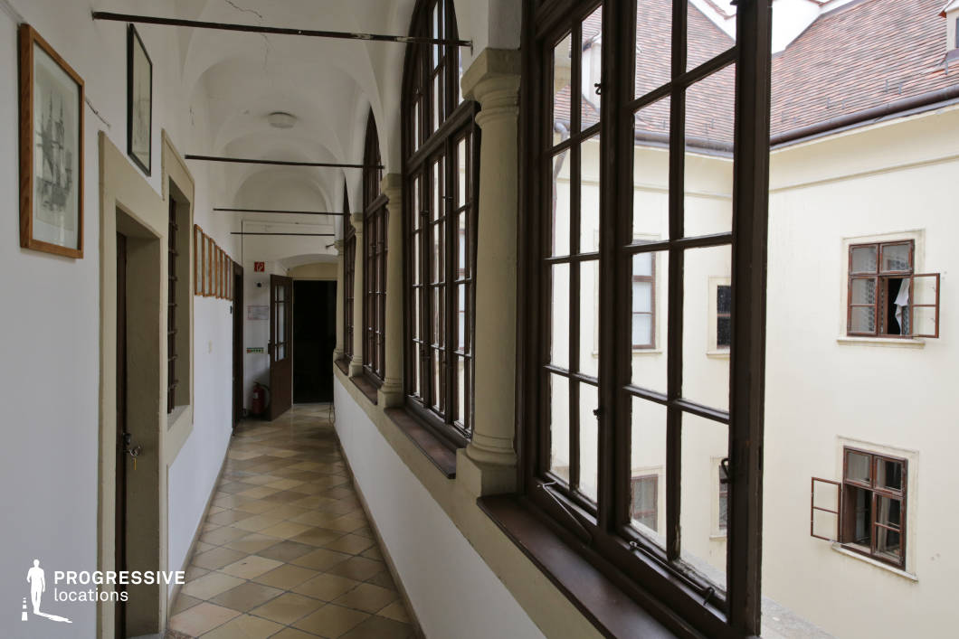 This image presents a narrow, arched corridor with a sequence of tall, multi-pane windows lining one side, opening onto a quiet inner courtyard. The space is defined by its vaulted white ceilings, thick plastered walls, and tiled floor in a subdued earth-tone palette, contributing to a serene and understated ambiance. The window frames and supporting columns exhibit traditional craftsmanship, while the visible wooden doors and framed wall art reinforce the building’s historical character. The symmetry of the corridor and the interplay between interior and exterior elements create a visually balanced composition. Ideal for period dramas, historical thrillers or introspective scenes, the location’s enclosed yet airy structure offers a controlled, cinematic setting with classical architectural elements.