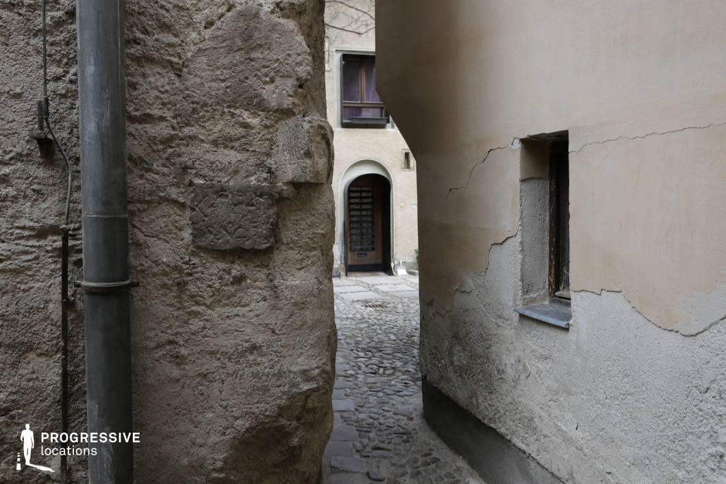This location presents a narrow stone alleyway framed by textured, weathered walls with visibly aged plaster and a muted, earthy color palette of beiges and greys. The rough stone facade on the left contrasts with the smoother, cracked surface of the opposing wall, highlighting the passage’s irregular geometry. The cobblestone pathway beneath adds to the old-world charm, leading the eye toward an arched doorway and shuttered window that suggest pre-modern architectural elements. The visual depth created by the passage’s tightness and the layered background enhances the cinematic appeal. Ideal for film productions requiring a historical European setting, this secluded urban corner is suitable for thrillers, dramas, or period films needing atmospheric, enclosed exterior locations.