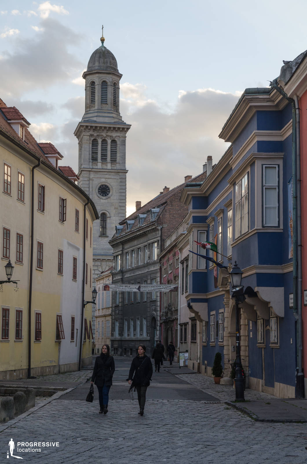 This image captures a narrow, winding cobblestone street framed by colorful, tightly-aligned historical buildings, leading the eye toward a tall, light stone church tower with classical architectural detailing and a domed top. The facades along the street exhibit a mix of pastel and bold tones—muted yellow, deep blue, and brick red—highlighted with detailed cornices and uniformly placed wooden-framed windows. The blue building on the right features a protruding bay window and stone column accents, adding visual depth to the scene. The street has a distinctly European old-town character with a human-scale layout, traditional streetlamps, and preserved architectural details from various eras. This location is ideal for filming period dramas, historical pieces, or character-driven scenes requiring an atmospheric, European setting with strong visual identity and compositional depth.