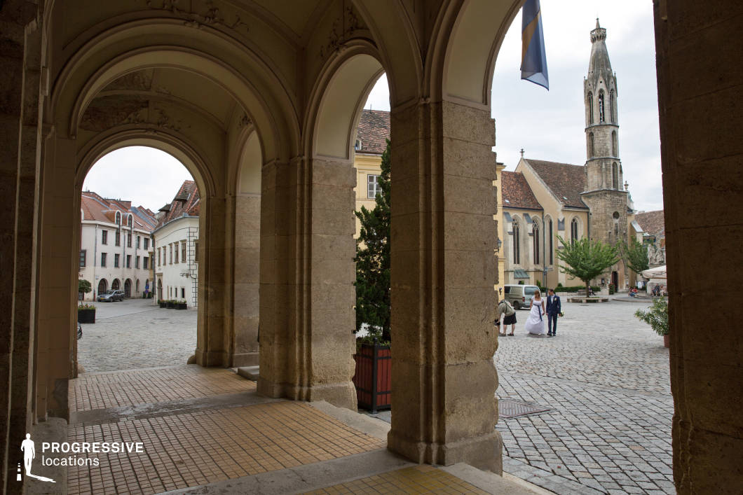 This location presents a refined combination of architectural elegance and urban charm, with a symmetrical arcade of large stone pillars and ornate vaulted ceilings framing the view of a traditional European town square. The covered passageway features warm-toned ceramic floor tiles and opens onto a spacious cobblestone plaza bordered by pastel-hued historic buildings and a prominent Gothic-style church with a tall, narrow bell tower. The structures showcase steep, tiled roofs and vertically oriented windows, reinforcing a classical, old-world aesthetic. The composition uses a central perspective with clear foreground-background depth, making it ideal for dynamic framing. The blend of architectural detail, open space, and strong visual lines makes this location highly adaptable for historical dramas, romantic scenes, and culturally grounded productions seeking an atmospheric, period-evoking setting.