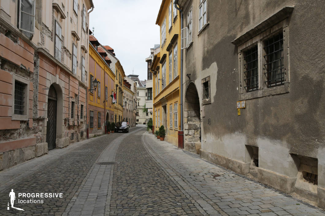 This location features a narrow, winding cobblestone street framed by multi-story historical buildings with a distinct patina of age. The facades are a mix of faded pastel tones and saturated ochre, showcasing exposed brickwork, chipped plaster, and iron-barred windows that evoke a weathered, centuries-old charm. The architectural style is predominantly classical with some baroque and vernacular elements, visible in the arched stone entrances, tall sash windows, and subtly decorative cornices. The spatial proportions are intimate, with tightly spaced buildings creating a visually enclosed corridor-like atmosphere. The gradual curve of the street leads the eye into depth, enhancing the compositional layering and allowing for dynamic camera movement. This alley provides a versatile backdrop for film projects requiring old European town settings, period authenticity, or a slightly gritty urban texture—suitable for dramas, historical pieces, thrillers, or atmospheric transitional scenes.