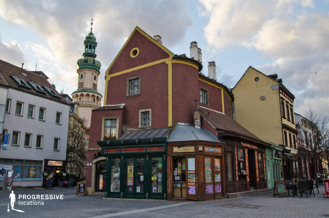 This location features a visually striking street corner defined by a bold, deep-red historic building accented with mustard yellow trim and a unique rounded extension. The architecture blends early urban European styles, with elements like steep tiled roofs, multi-pane windows, and contrasting materials such as textured plaster façades and detailed wooden storefronts. A small currency exchange booth and multiple shopfronts line the base, adding functional depth and vintage charm. Adjacent buildings exhibit a mixture of renovated and weathered exteriors, including pastel and earth-toned façades, providing a layered historical context. The street is paved with uniformly laid cobblestones, suggesting pedestrian accessibility and urban intimacy. The vertical presence of a baroque-style tower in the background enhances the sense of place and adds architectural complexity. This setting is ideal for filming period dramas, European-themed scenes, or character-driven urban narratives due to its textured surfaces, compact scale, and rich atmospheric presence.