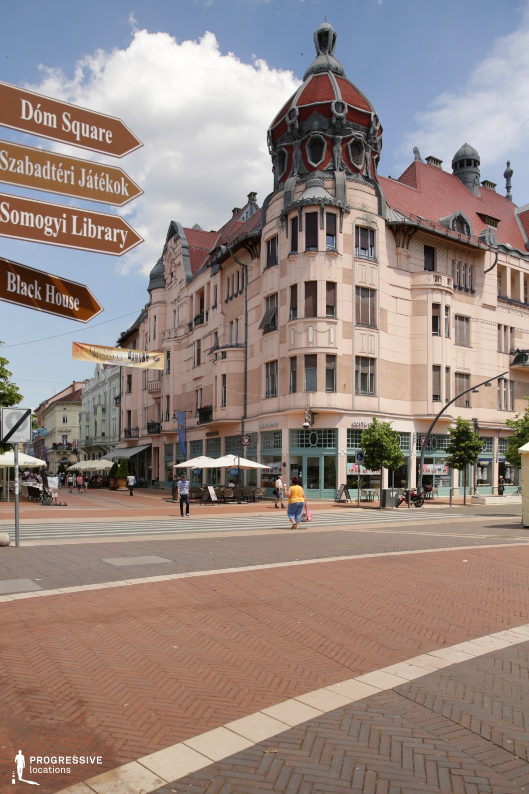 This visually distinctive location is defined by an elaborate corner building in Art Nouveau style, characterized by ornate turrets, curved balconies, and a decorative roof structure with intricate stone and ironwork. The pastel-toned façade incorporates soft pink hues complemented by red roof tiles and elegant, rounded window details. The building anchors a lively pedestrian square paved in patterned brickwork, with clear walkways, mature trees in planters, and visible café terraces along the ground floor. Directional signage and urban furniture contribute to the organized yet welcoming character of the space. The combination of historical architecture, richly textured materials, and open pedestrian access makes this an ideal setting for period films, romantic comedies, cultural documentaries, or fashion productions seeking a distinctive European urban aesthetic with stylistic flair.