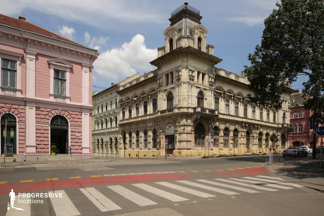 This urban street corner features richly ornamented 19th-century architecture, dominated by a grand, multi-story historic building with a central tower structure and elaborate window framing. The facades are detailed with classical elements such as pilasters, arched windows, and cornices, presenting a blend of neoclassical and eclectic styles. The color palette transitions from soft pinks to weathered creams and beige, contributing to a romantic, old-European city atmosphere. The buildings display significant verticality with two to three stories, and their proportions enhance the enclosed yet spacious character of the intersection. The crosswalk and street markings introduce linear geometric elements, further defining the setting’s compositional clarity. This location is ideal for filming period dramas, urban explorations, or atmospheric city scenes, thanks to its architectural depth, visual symmetry, and strong historical ambiance.