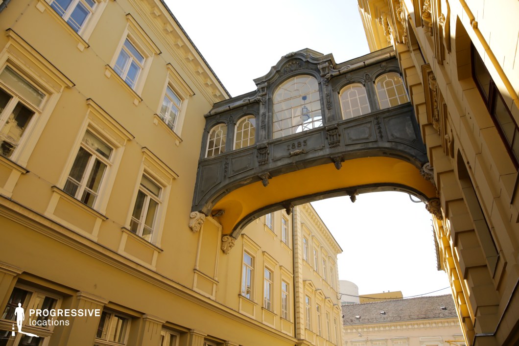 This narrow urban passageway is visually defined by an ornate, covered skybridge that connects two classical-style buildings. The bridge, dated 1883, features richly detailed ironwork and large arched windows, creating a visually striking architectural element that contrasts with the smooth, pastel-yellow façades surrounding it. The buildings themselves display symmetrical window arrangements, subtle cornices, and decorative stone accents typical of late 19th-century European architecture. The upward perspective emphasizes the verticality of the walls and the central prominence of the skybridge, while the golden-yellow tones throughout the scene provide a cohesive, warm atmosphere. This location offers a unique visual character ideal for period dramas, historical documentaries, or scenes requiring a distinctive European alleyway with architectural depth and a sense of enclosure.