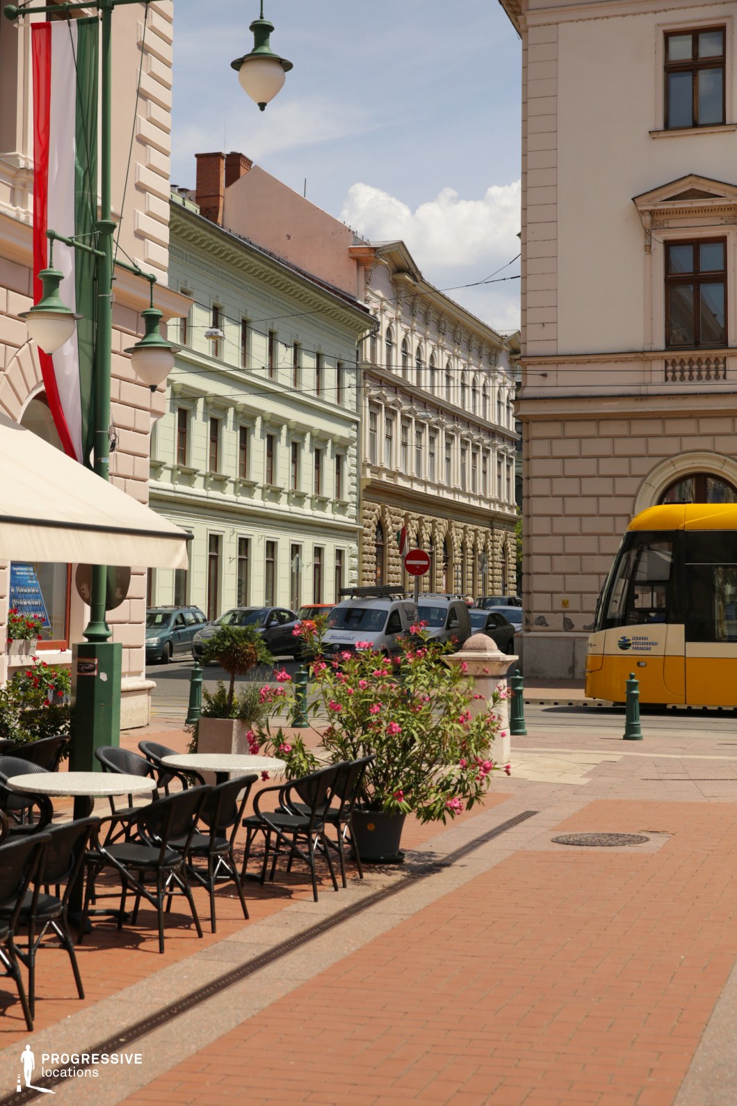 This image captures a charming, structured urban corner characterized by historic European architecture with homogeneous neoclassical facades in pastel shades of green and beige. The buildings span multiple stories and feature symmetrical window arrangements, cornices, and ornamental details that contribute to a refined, cohesive cityscape. The foreground is dominated by a paved pedestrian zone with an outdoor café terrace, marked by metal-framed round tables and chairs aligned in neat rows. A yellow tram adds a dynamic, functional element along the embedded rail line, contrasting with the otherwise static environment. The composition, taken from a slightly low eye-level angle, emphasizes both depth and linear perspective as the street narrows into the background. The scene offers a harmonious blend of urban elegance and public infrastructure, making it an ideal location for filming period pieces, romantic scenes, or lifestyle-oriented commercials within a European city setting.