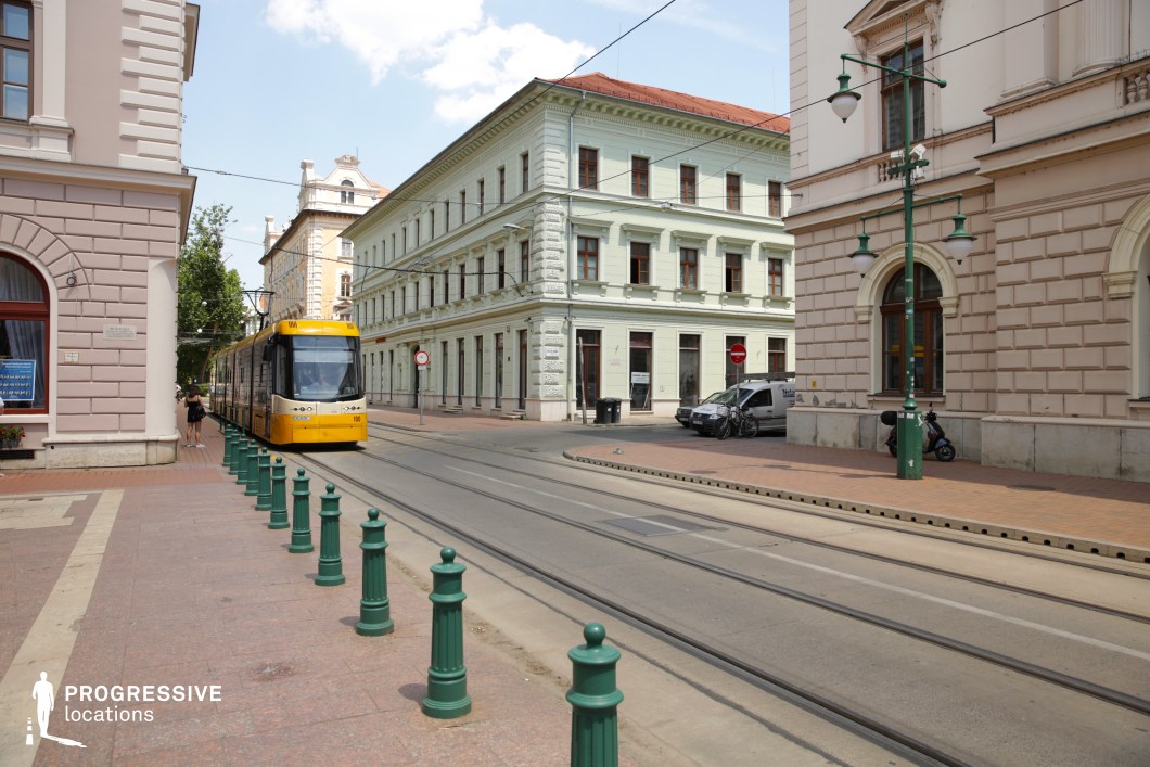 This urban location features a clean, symmetrical tram corridor flanked by stately neoclassical buildings with finely detailed façades. The architecture presents a refined palette of pale tones, including soft cream and mint green, with rhythmic window arrangements and decorative cornices that enhance the formal elegance. The tram tracks run centrally through the image, accompanied by a row of green cast-iron bollards and period-style street lamps, reinforcing the historical cityscape ambiance. The wide, paved sidewalks and corner structures contribute to a spacious and orderly composition. This location is highly suitable for contemporary urban scenes, historical reconstructions, or European city atmospheres in genres such as drama, romance, or political thriller.