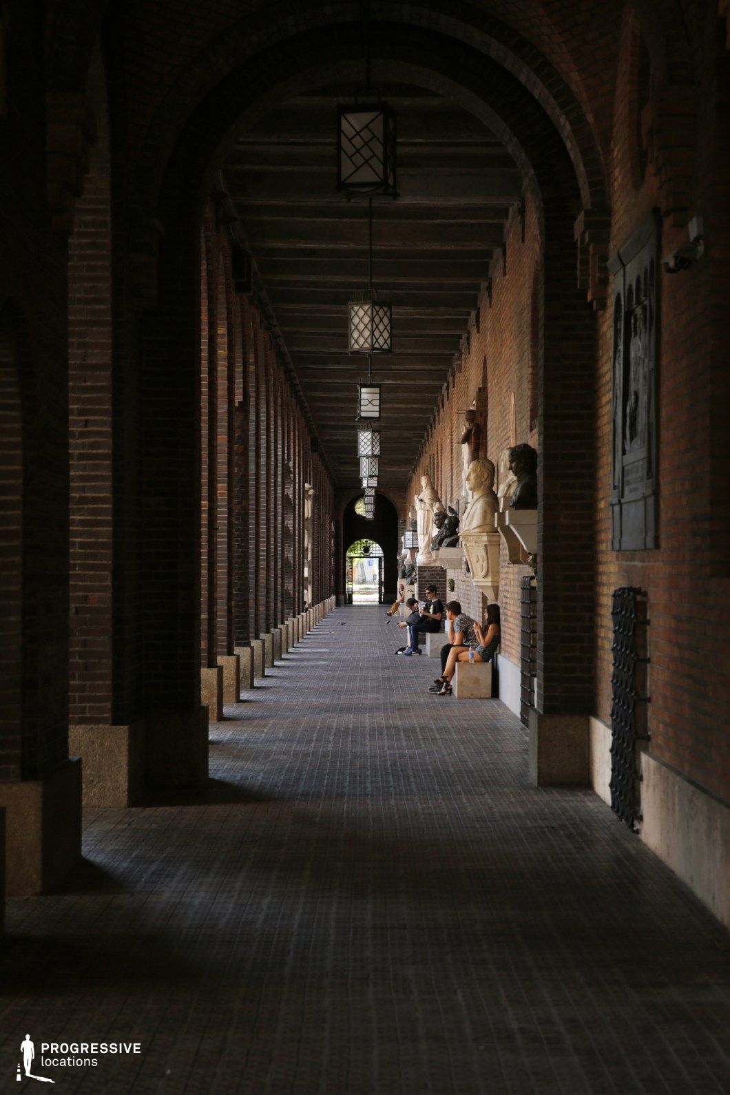 This location features a long, covered arcade defined by deep red brick walls, repeating rounded arches, and symmetrical hanging lanterns that establish a strong sense of rhythm and depth. One side of the corridor is lined with sculpted busts and reliefs embedded into the wall, adding an academic or memorial character to the space. The floor is tiled in a grid-like pattern, enhancing the corridor’s linear geometry and further guiding the eye through the perspective. The architecture is classical-influenced with a somber, introspective tone created by the dim natural light filtering in from the arch openings. This enclosed, monument-like setting is ideal for period dramas, historical productions, or introspective scenes requiring formal, reverent, or institutional atmospheres.