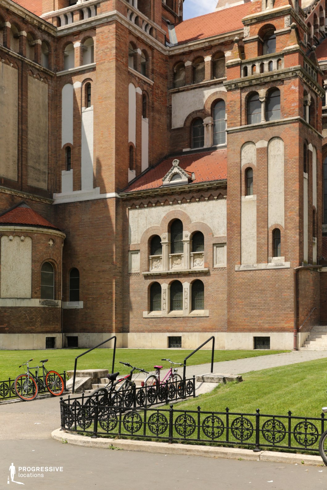 This location highlights a richly detailed Romanesque Revival building façade, featuring tall brick towers, rounded arched windows, and stone decorative elements that convey a sense of monumentality and historical gravitas. The red and beige tones of the masonry are complemented by detailed cornices and carved stone panels, adding texture and visual interest. The small courtyard in front is framed by ornamental black fencing and gently sloping green lawns, providing a clean and symmetrical foreground. The space offers a structured, almost cloister-like atmosphere, ideal for period films, historical dramas, or scenes requiring religious, academic, or culturally significant settings with an enclosed, contemplative feel.