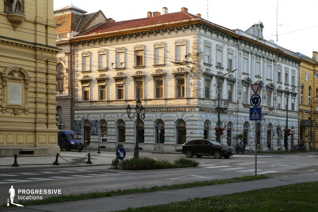 This urban location captures a set of late 19th-century and early 20th-century corner buildings with richly ornamented façades and neoclassical detailing, including arched windows, pilasters, cornices, and decorative sculptures. The buildings form a compact, formal streetscape with a variety of pale hues—creamy yellow and white—contrasting subtly against the red-tiled roofs. The intersection is structured with wide pedestrian paths, cast-iron lamp posts, and controlled roadways, creating a visually ordered and historic city center atmosphere. This location is well-suited for period dramas, political thrillers, or any genre requiring elegant European architectural settings with layered spatial composition and a sense of civic formality.