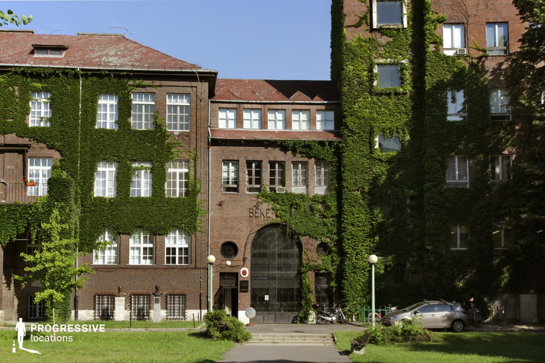 This location features a large, symmetrical red-brick institutional building partially overgrown with dense ivy, creating a striking visual contrast between natural and architectural elements. The structure is characterized by its tall rectangular windows arranged in a rhythmic grid, a grand arched entryway with ornamental glasswork, and steeply pitched tiled roofs. The use of traditional materials like brick and stone adds a robust, historic atmosphere, while the ivy softens the appearance and adds texture. The building’s proportions suggest a mid-20th-century institutional or educational function, with an expansive courtyard in front that offers visual depth and framing options for wide or frontal shots. This visually layered and architecturally rich setting is well-suited for historical, academic, or dramatic genres, particularly scenes requiring an evocative institutional backdrop or classic urban courtyard.
