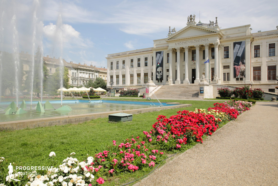 This location showcases an imposing neoclassical museum building, defined by its grand columned portico, triangular pediment, and symmetrical white façade adorned with ornate reliefs and statues. The wide stone staircase leads up to the entrance, enhancing the sense of formality and grandeur. In the foreground, a landscaped plaza features a geometric fountain with vertical water jets and manicured flowerbeds lined with vibrant red, pink, and white blooms. The surrounding lawn and walking paths create a refined yet accessible public space. The architectural style evokes institutional elegance, while the open layout provides ample depth and layering for long shots or dynamic camera movement. This location is ideal for cultural, academic, or governmental scenes in films and series, and works well for genres including drama, historical fiction, or contemporary productions seeking a prestigious urban backdrop.