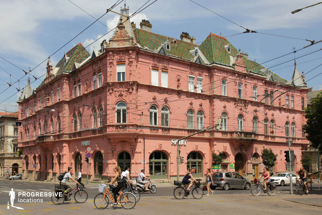 This eye-catching urban location features a richly ornamented Art Nouveau-style corner building painted in a vibrant pink hue, immediately commanding visual attention. The façade is detailed with intricate stucco work, arched windows, and decorative elements underlining the building's historical elegance. A steeply pitched roof clad in green patterned tiles and punctuated by small dormers crowns the structure, enhancing its picturesque appearance. The architectural symmetry and prominent corner position give it a strong spatial presence within the intersection. This location is ideal for romantic, historical, or European-set productions and is especially effective for scenes requiring a distinctive, visually striking backdrop with a nostalgic or whimsical ambiance.
