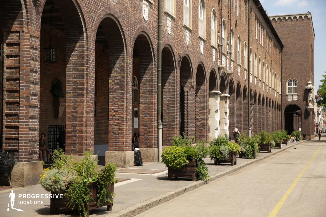 This visually commanding location features a long, continuous colonnade of rounded brick archways forming the base of a red brick institutional building. The repetitive rhythm of the arches, constructed in dark-hued brick with stone accents, creates a strong linear perspective and monumental effect. Above the arcade, regularly spaced windows are interspersed with stone crests and sculptural reliefs, contributing to a formal, almost fortress-like character. The massive scale is emphasized by the building’s sheer length and the elevated positioning of its architectural details. Large planters with dense greenery soften the austere façade and add a layer of texture along the wide, lightly paved pedestrian lane. The combination of historic institutional architecture and bold structural geometry makes this an excellent setting for political dramas, historical narratives, or stylized fashion shoots that require a backdrop with gravitas and architectural continuity.