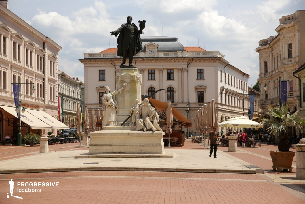 This image captures a spacious, paved town square dominated by a multi-figure stone monument at its center, featuring a commanding bronze statue atop a classical pedestal. The surrounding architecture consists of elegant, historic buildings with neoclassical and baroque details, including arched windows, pilasters, and ornate cornices in soft pastel tones. The central building in the background reinforces the symmetrical composition with its balanced façade and clock detail. Large umbrellas and aligned outdoor seating areas suggest commercial or hospitality use, enhancing the square's function as a civic and social hub. The reddish-brown paver stones and light stone pathways provide visual contrast and structure, guiding movement through the open space. The clear sightlines, central monument, and rich architectural backdrop make this location highly suitable for filming historical dramas, period pieces, political thrillers, or promotional content requiring a refined European city ambiance.