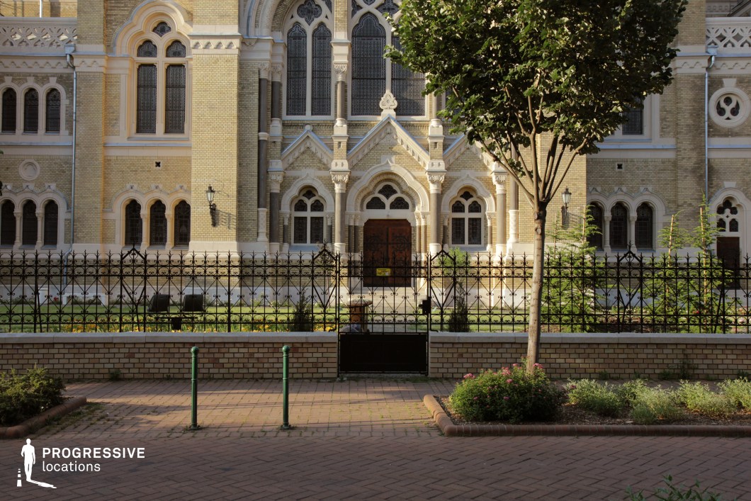 The image presents a detailed frontal view of a historic building entrance characterized by refined neo-gothic architecture. The symmetrical composition centers on a prominent triple-arched portal framed by decorative stonework and pointed arch windows with intricate tracery. The façade is constructed from light yellow brick, accented with white stone elements that highlight architectural features such as columns, arches, and window frames. Wrought iron fencing with ornate motifs encloses a meticulously maintained garden, enhancing the building’s dignified presence. The overall geometry of the scene is balanced and formal, with vertical lines dominating the composition. This location is ideal for period films, historical dramas, or scenes requiring a stately institutional backdrop. Its classic design and clear architectural identity make it a versatile setting for both cinematic establishing shots and dramatic entrances.
