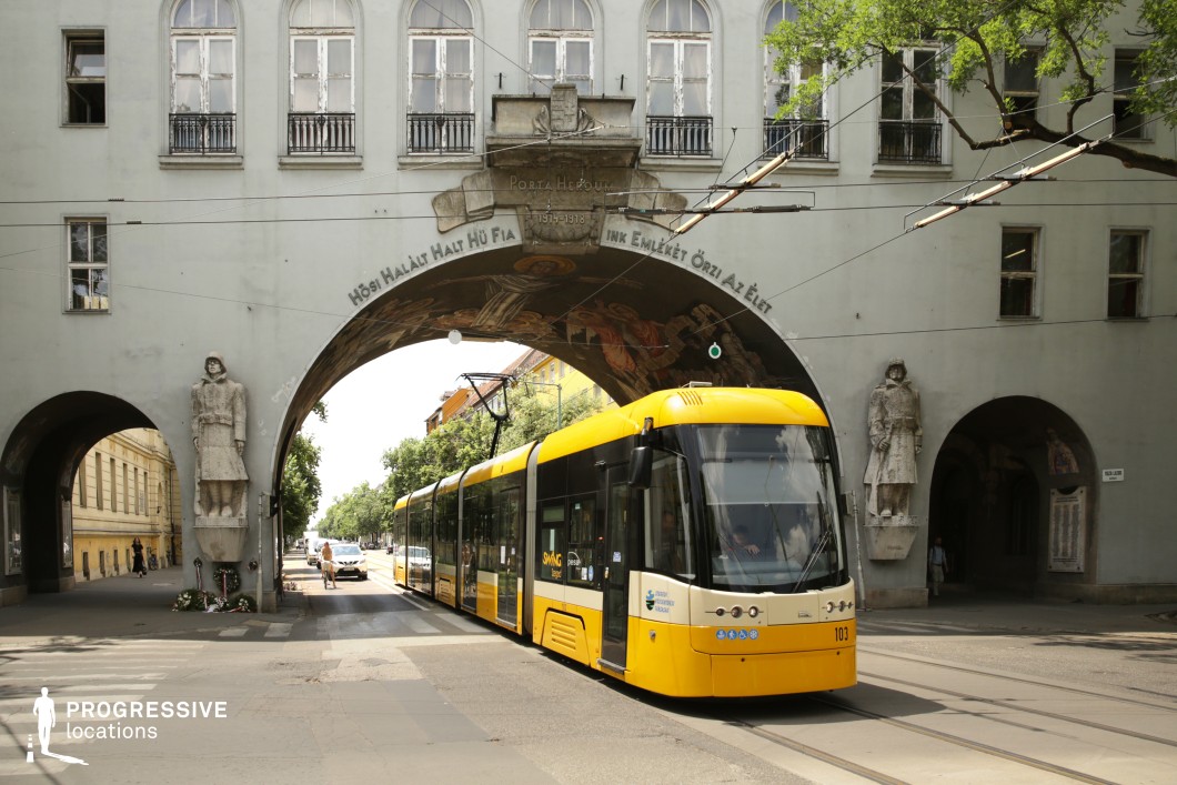 This urban location features a distinctive architectural ensemble where a broad streetcar route passes directly beneath a monumental archway integrated into a classical-style building façade. The arch is framed by sculpted stone statues and decorated with a mural on its vaulted underside, adding historic and artistic character. The muted gray exterior of the structure contrasts with the vivid yellow tram, emphasizing the interplay between traditional and contemporary urban elements. The symmetrical composition, combined with the tram tracks leading through the arch, creates strong linear geometry and visual depth. This location is ideal for urban scenes with historical undertones, and suits drama, period pieces, or productions emphasizing civic or metropolitan environments.