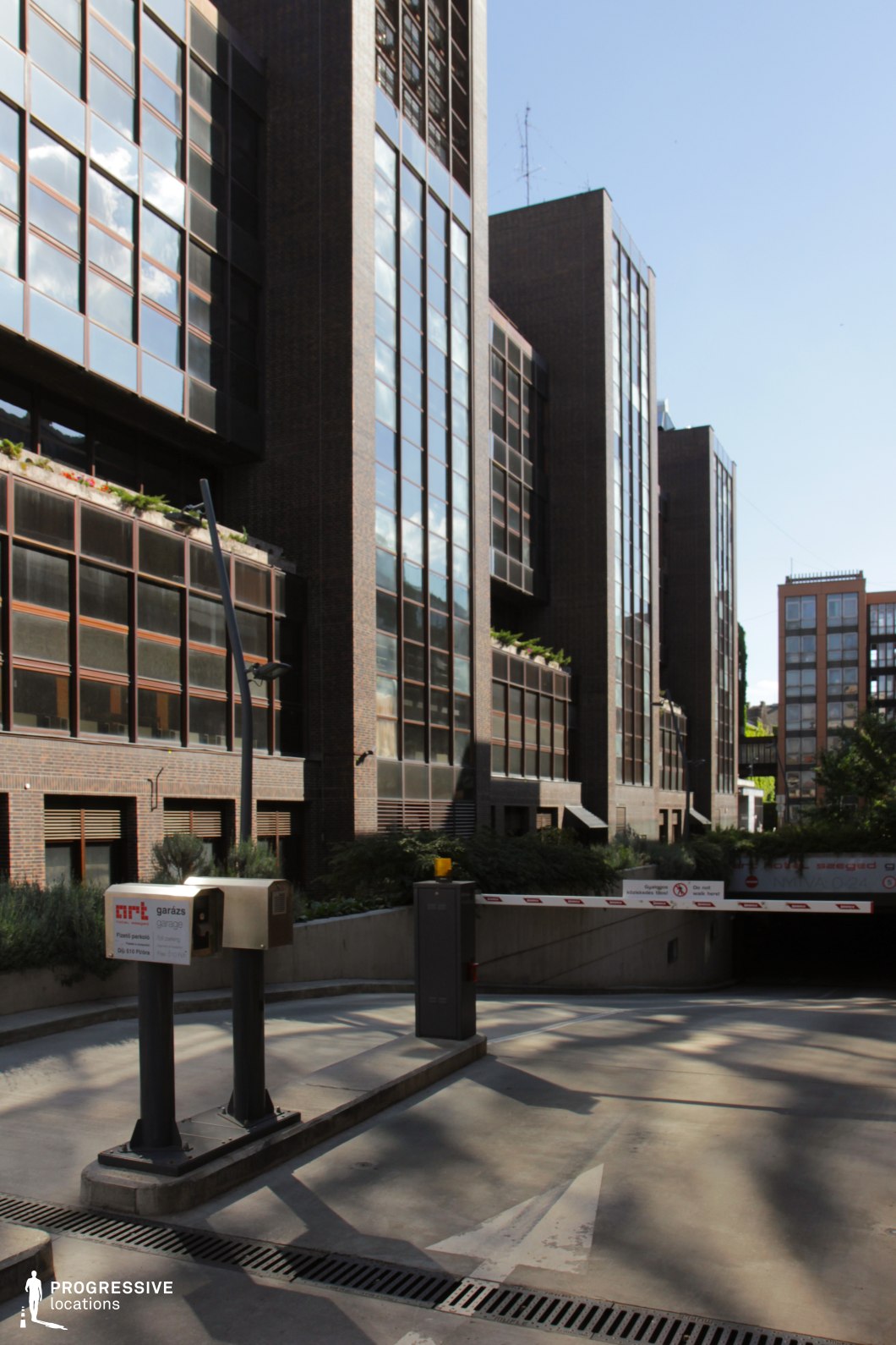 This location showcases a sleek, modern office complex characterized by tall, narrow glass façades framed with dark brown brick cladding. The strong vertical lines of the buildings are accentuated by the rhythmic repetition of windows, creating a corporate and structured visual language. In the foreground, a clearly defined underground garage entrance is visible, marked by a barrier gate system and directional road markings, adding a utilitarian and functional layer to the setting. The buildings’ proportions and reflective surfaces lend the scene a distinctly urban and contemporary atmosphere. This site is highly suitable for corporate, thriller, or action genres, and works well for scenes involving office environments, surveillance sequences, or underground transitions.