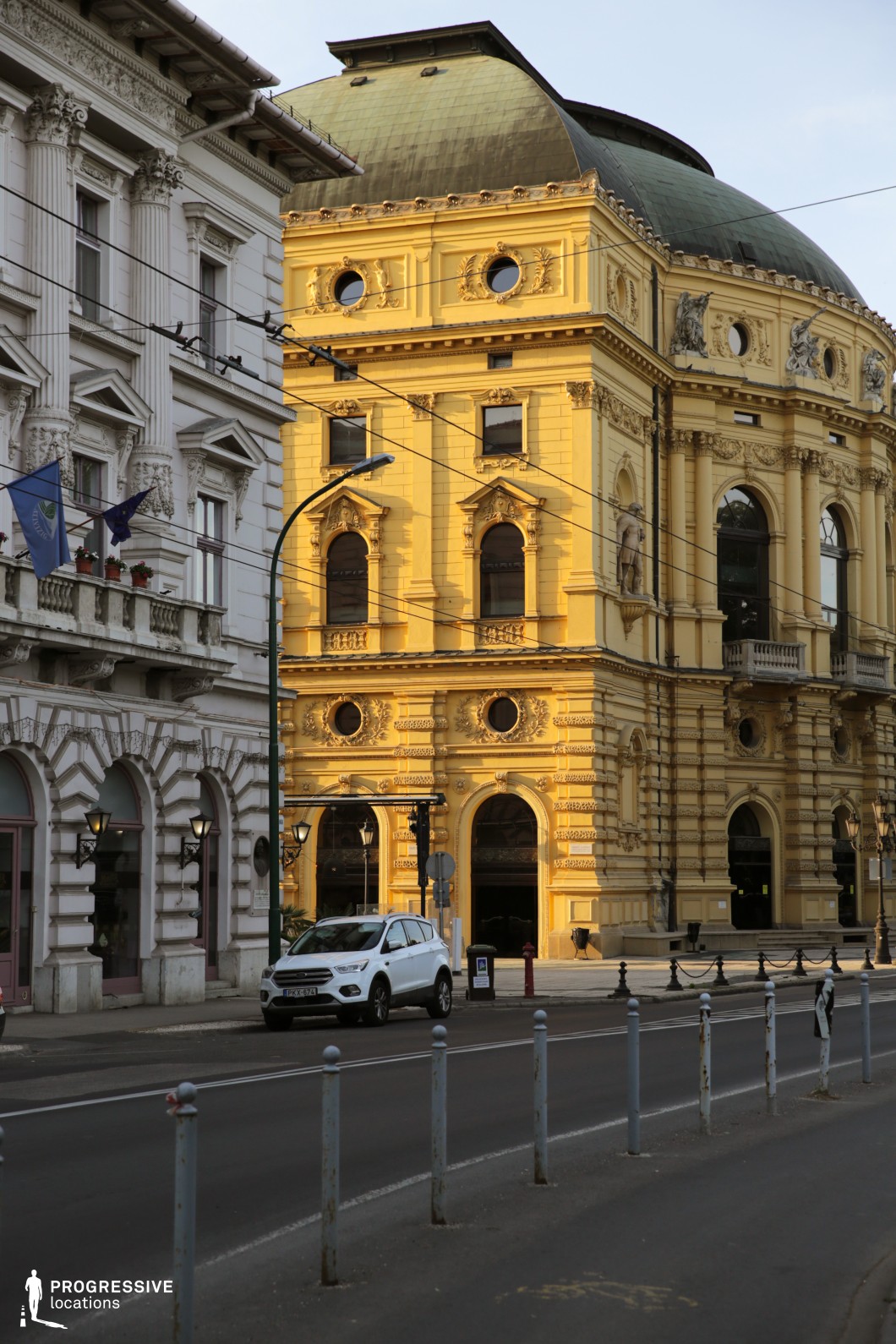 The image presents an impressive urban streetscape dominated by a richly detailed neoclassical theater building with a yellow facade and green oxidized copper dome. The architecture is marked by high arched windows, elaborate cornices, sculpted reliefs, and pilasters, contributing to a stately and historic atmosphere. The building spans multiple floors, with monumental scale and strong vertical emphasis, enhanced by deep window recesses and ornamental window crowns. Adjacent structures feature classical motifs and white stone cladding, creating a strong visual contrast that emphasizes the theater's vibrant color and ornate detail. The camera angle, positioned at street level, accentuates the verticality of the scene while capturing the intersection and open road in the foreground, adding a sense of urban dynamism. This location is ideal for period films, dramas, or upscale urban scenes, and is particularly suitable for productions requiring a sophisticated architectural backdrop or a city center with European character.
