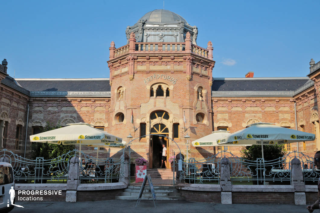 This ornate building features a richly detailed Art Nouveau facade constructed from warm-toned brick, characterized by symmetrical arched windows, decorative stonework, and a central rotunda crowned by an intricate dome. The foreground is defined by an elegant wrought-iron fence and stone posts enclosing a small terrace area, complemented by large branded parasols and a formal staircase leading to the grand arched entrance. Architectural embellishments such as pilasters, friezes, and curved balconies lend the structure a regal yet inviting presence. The composition is balanced and frontal, enhancing the building’s architectural symmetry. This location is highly versatile for filming, particularly suited for historical dramas, romantic narratives, or cultural documentaries, offering a distinctive Central European backdrop with heritage-rich atmosphere and detailed period architecture.