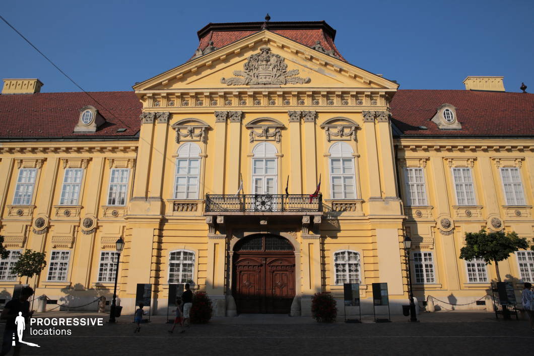 This stately Baroque-style palace features a symmetrical facade dominated by a central pediment adorned with elaborate sculptural reliefs and a crest. The bright yellow plastered exterior, combined with classical white-framed windows and ornate stone details, evokes an elegant and historical ambiance. The richly carved wooden double doors serve as a focal entry point beneath a prominent balcony with wrought iron railing and a central ornamental clock. The building is flanked by repetitive pilasters and evenly spaced vertical windows, contributing to a rhythmically structured and formal appearance. The mansard-style roof, covered with red tiles, adds a distinctive silhouette to the structure. The spacious cobblestone forecourt enhances the grandeur of the building and allows for broad camera angles. This location is ideal for period films, historical dramas, or any genre requiring a regal or institutional backdrop, such as political thrillers or formal ceremonial scenes.