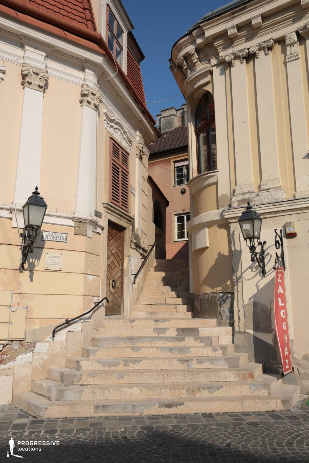 This location features a narrow stone stairway nestled between two ornate, classical-style buildings, forming a visually enclosed and vertically layered urban space. The worn limestone steps and aged façades evoke a strong sense of architectural patina, with chipped plaster, richly textured surfaces, and detailed stucco ornamentation. The buildings display baroque and eclectic elements such as pilasters, cornices, and curved bay windows, while the muted cream and ochre color palette enhances the old-world atmosphere. Wrought iron lanterns and robust wooden shutters contribute to the period charm. The ascending perspective leads the eye inward, creating a natural depth and visual rhythm. This location is ideal for period dramas, historical reconstructions, or atmospheric urban scenes, particularly those requiring a sense of timeworn authenticity or confined, vertically structured environments.
