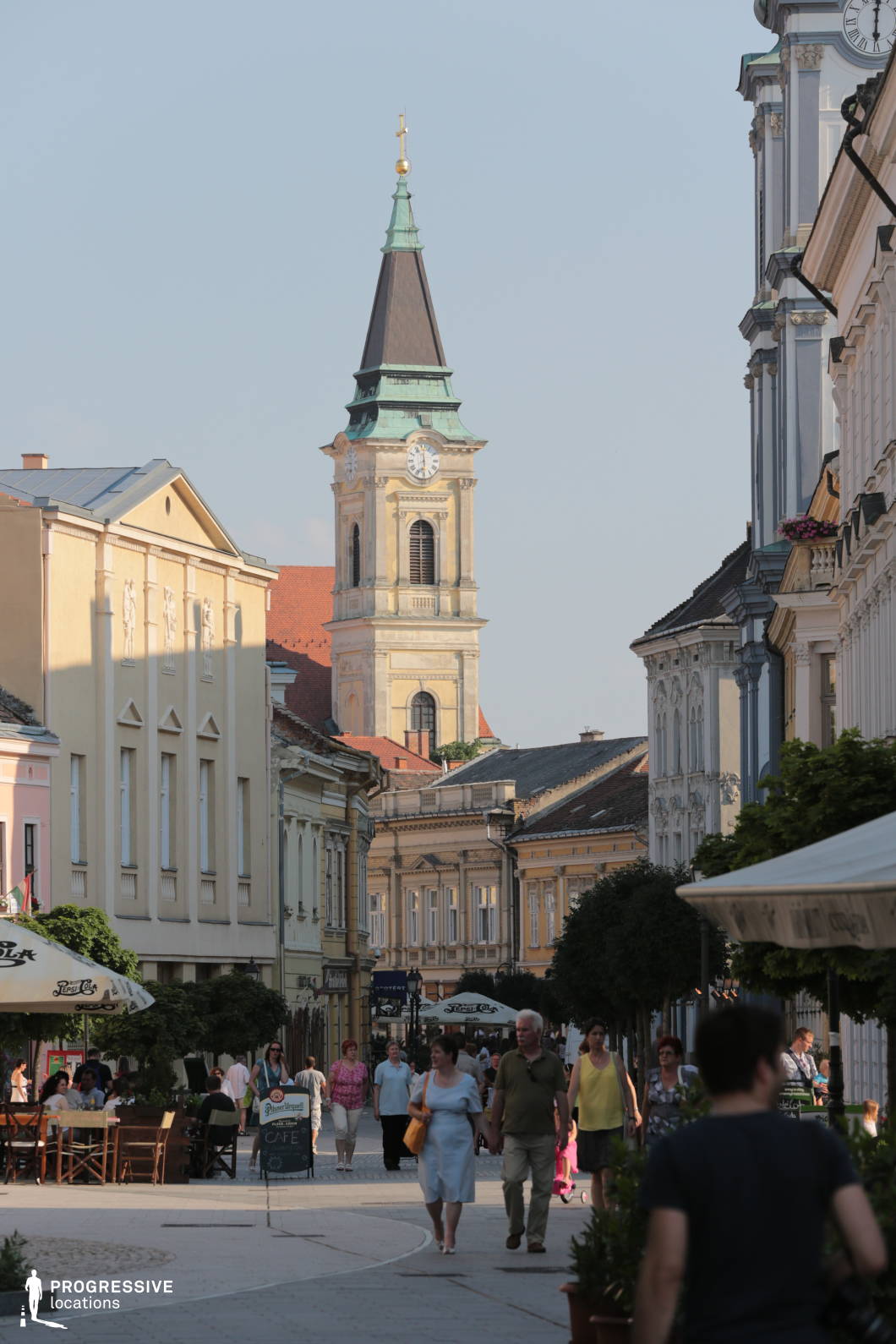 This image captures a charming, architecturally cohesive pedestrian street lined with pastel-toned historic buildings, most featuring ornate window moldings, classical cornices, and Baroque or Neoclassical elements. The prominent church tower with a clock face and a distinctively green patinated roof anchors the visual composition, rising above the low-rise urban fabric. The buildings are closely set, creating an intimate streetscape ideal for walkability and outdoor seating areas, supported by wide stone-paved walkways. This location's elegant facades and symmetrical streetscape make it highly adaptable for filming period dramas, romantic scenes, or upscale commercial shoots requiring a European small-town atmosphere.