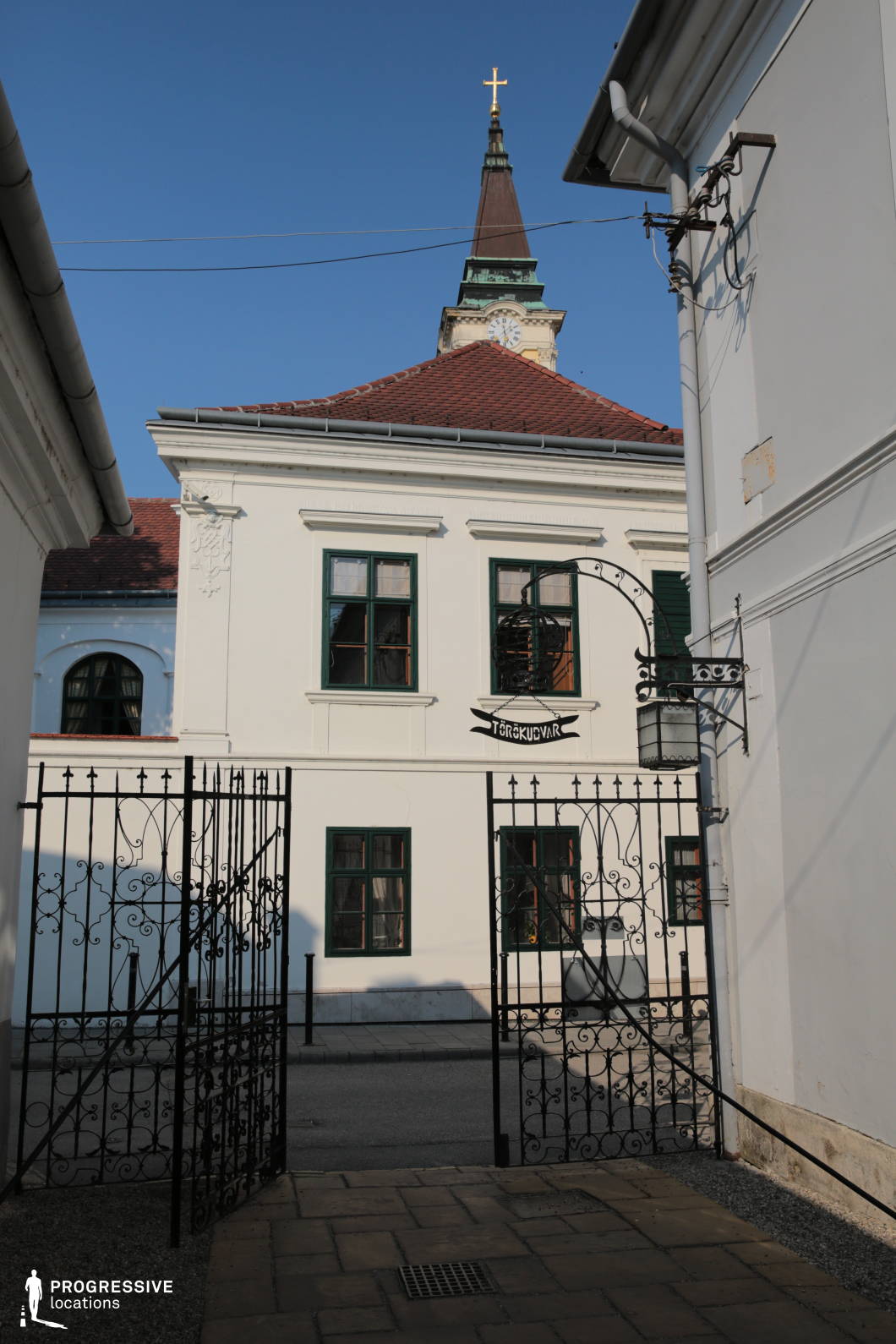 This location presents a narrow, enclosed courtyard framed by white, classicist-style buildings featuring simple, geometric façades with dark-framed rectangular windows. The architectural lines are clean and symmetrical, and the white plastered walls contrast with the red tile roofing. A dominant vertical element rises in the background—a church tower with a steeply pitched spire, clock face, and cross—adding historical depth and verticality to the composition. The ornate wrought-iron gate in the foreground introduces delicate decorative detailing and defines a clear transitional threshold into the space. The perspective is frontal and slightly low-angle, emphasizing the symmetry and enhancing the sense of enclosure. This visually structured and historically resonant location is ideal for period dramas, contemplative urban scenes, or narratives requiring formal architectural elegance and a sense of spatial intimacy.