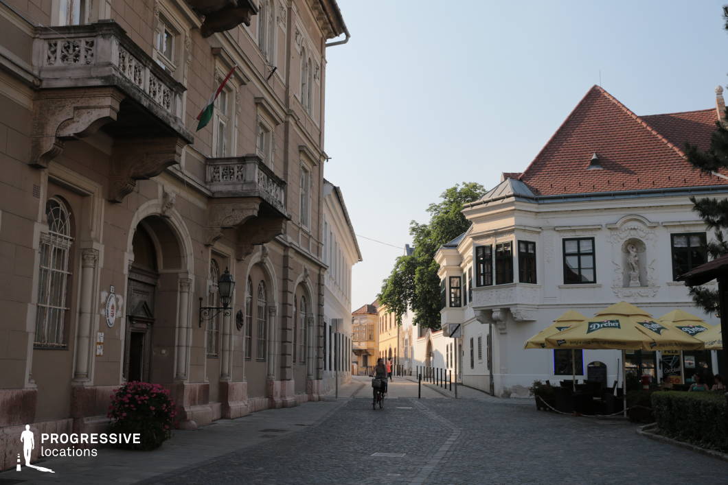 This picturesque urban scene showcases a narrow cobblestone street flanked by well-preserved historical architecture, including Baroque and Neoclassical styles. The left side features a beige-toned, arched façade with decorative balconies, intricate stonework, and wrought iron elements, evoking a formal, civic character. Opposite, a white corner building with ornate stucco detailing, bay windows, and a steep red-tiled roof adds architectural contrast and charm. The visual depth created by the winding street, combined with the subtle rhythm of the windows and decorative façades, reinforces the location’s timeless European atmosphere. This setting is ideal for period dramas, romance films, or cultural documentaries, offering a refined and elegant backdrop for scenes requiring a historical or intimate old-town environment.