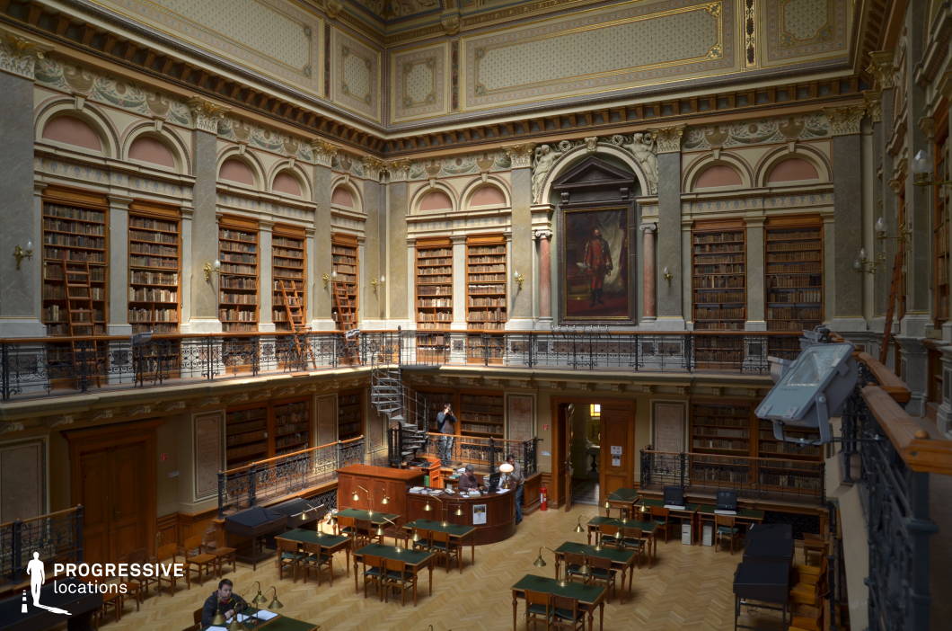 This grand Baroque-style university library hall exudes elegance and historical prestige, characterized by its richly decorated high ceilings, intricate moldings, and opulent architectural details. The room is encased by towering bookshelves filled with antique volumes, accessible via wooden ladders, reinforcing the classical academic ambiance. The upper-level balcony, lined with ornate wrought iron railings, adds depth and visual complexity to the space. A large, regal portrait framed in a lavish arch dominates the central wall, further emphasizing the hall’s historical and cultural significance. The parquet flooring and dark wood furnishings, combined with warm lighting, create a refined and scholarly atmosphere, making this location ideal for period dramas, intellectual-themed productions, and historical documentaries.