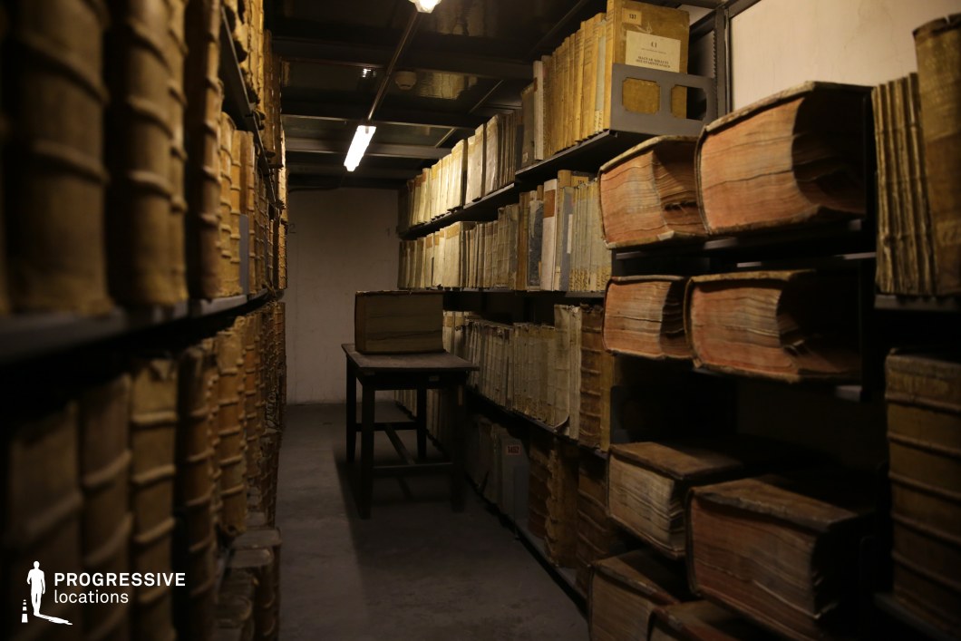  This dimly lit library archive exudes a sense of mystery and historical significance, with towering metal shelves stacked with ancient, leather-bound tomes and oversized manuscripts. The narrow aisle, flanked by aged books in various states of wear, leads toward a simple wooden table, reinforcing the space’s archival purpose. The subdued lighting, provided by a single fluorescent fixture, casts soft shadows that accentuate the worn textures of the book spines and the rough, unadorned concrete floor. The atmosphere is both intimate and solemn, making this location ideal for historical research-themed productions, mystery narratives, or period settings that require an air of secrecy and discovery.