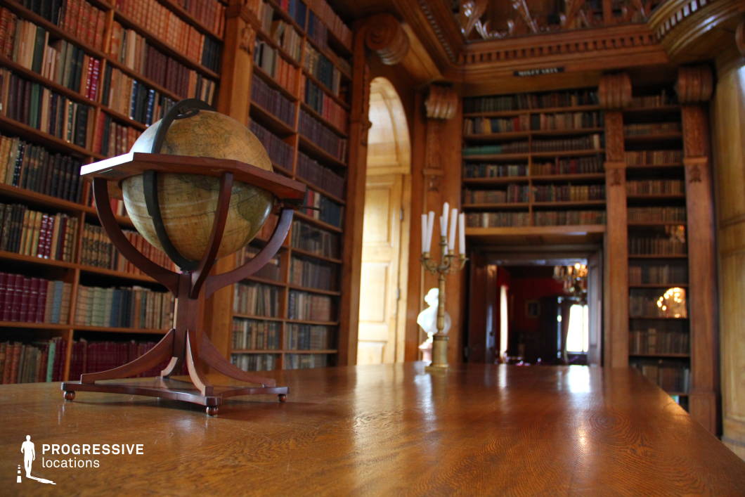 This atmospheric library scene features a vintage globe resting on a polished wooden table, surrounded by towering bookshelves filled with antique volumes. The warm, rich wood tones of the shelves and furniture contribute to the room’s intellectual charm. Intricate details, such as the candelabra and the architectural carvings along the ceiling, further enhance the historical and scholarly ambiance. A door at the far end of the room leads to a deeper section, where soft light from a chandelier highlights the luxurious setting. This refined, classic environment is ideal for period dramas, historical productions, or scenes that require an aura of scholarly prestige and timeless elegance.