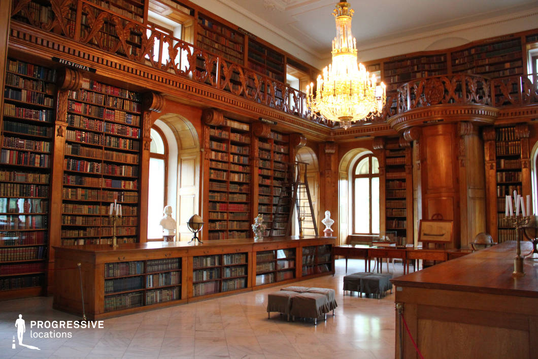 This lavish palace library embodies classical sophistication, featuring soaring wooden bookshelves filled with antique volumes, accessible by ladders and a gracefully curved mezzanine balcony. A grand crystal chandelier dominates the ceiling, casting warm, golden light that enhances the rich wood tones and polished marble flooring. Tall arched windows allow natural light to stream in, illuminating the elegant busts, globes, and vintage furnishings that decorate the space. The intricate wood carvings and refined architectural details evoke an atmosphere of aristocratic grandeur, making this setting ideal for period films, literary adaptations, or historical productions that require an opulent intellectual backdrop.