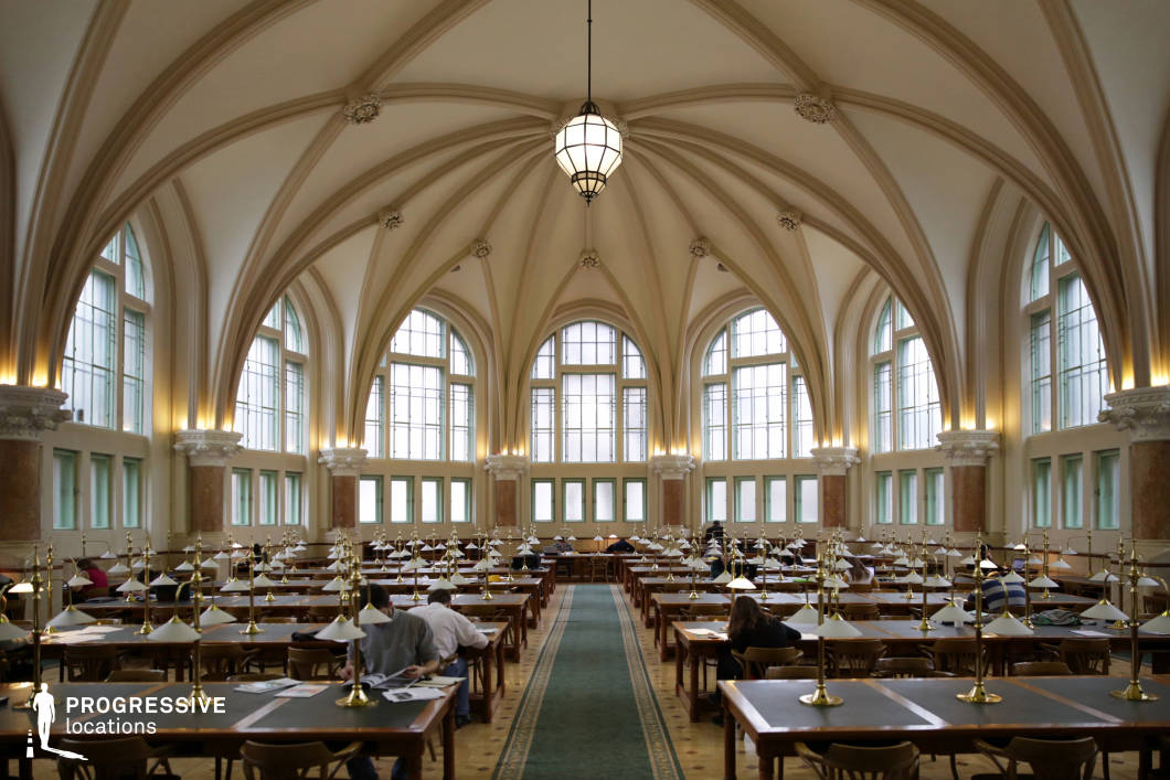 This Gothic-style university library hall features an expansive vaulted ceiling with elegantly ribbed arches that create a dramatic and imposing atmosphere. The high, pointed arches and intricate detailing evoke a sense of history and grandeur, while the large arched windows allow ample natural light to filter in, casting soft illumination across the space. The symmetrical layout enhances the room’s architectural harmony, with long wooden tables arranged in neat rows, each adorned with brass reading lamps that add a warm, vintage aesthetic. The polished wood flooring and muted cream-colored walls contribute to a refined, academic ambiance. This setting is ideal for capturing scholarly, historical, or period-piece productions, emphasizing intellectual depth and classical sophistication.
